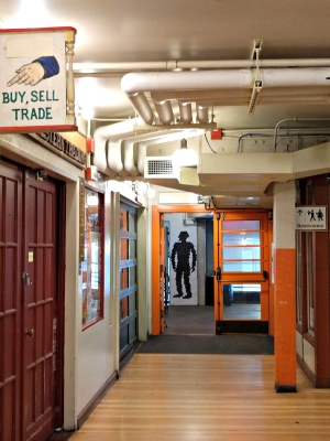 View down hallway at Pike Place Market; "Buy Sell Trade" sign is visible in foreground, while a door with cartoon man's silhouette is visible in background.