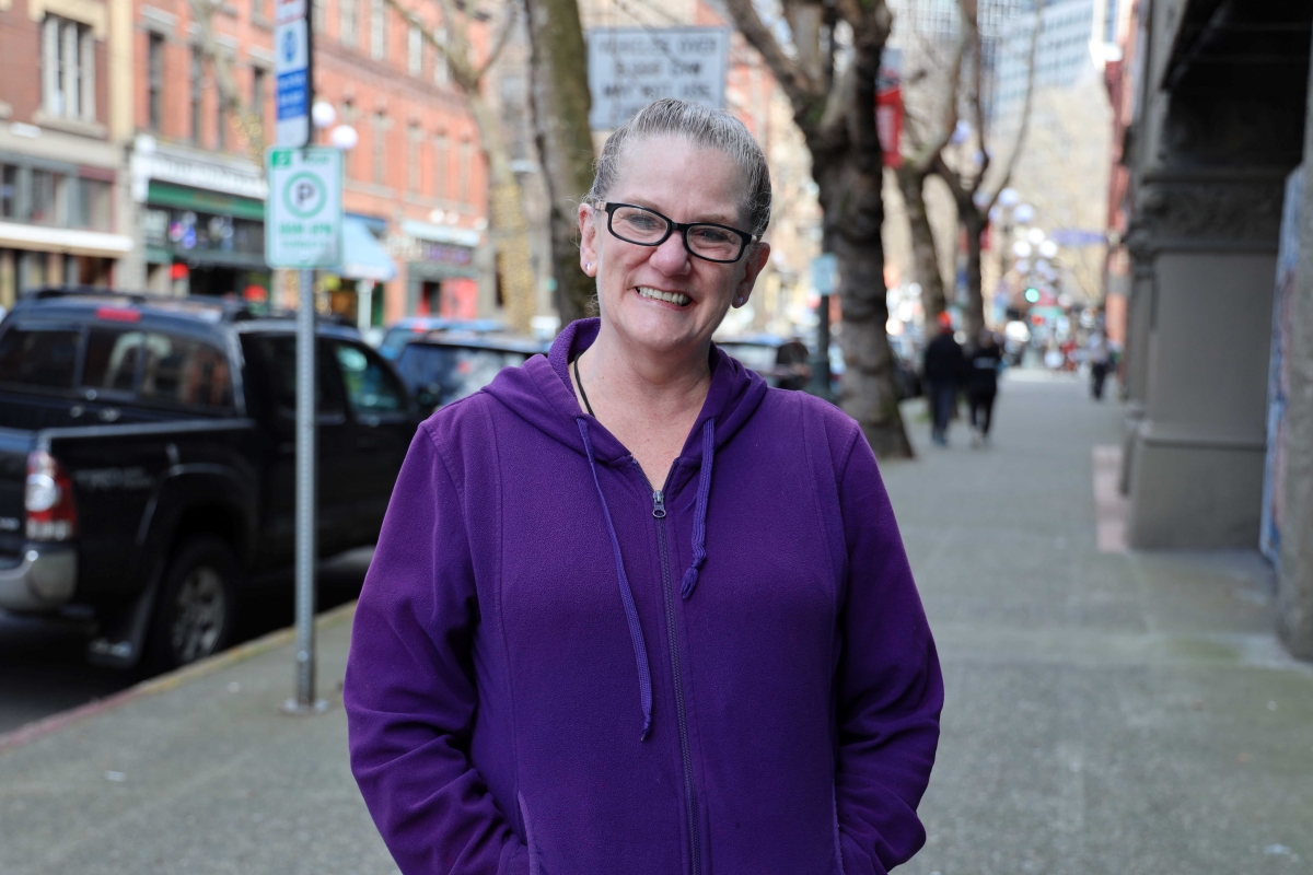 A middle-aged woman in a purple hoodie with glasses facing the camera on a tree-lined brick sidewalk.