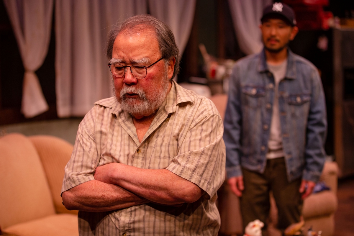 An older Asian man with a gray beard and glasses facing the audience with his arms crossed, while another actor is out of focus in the background.