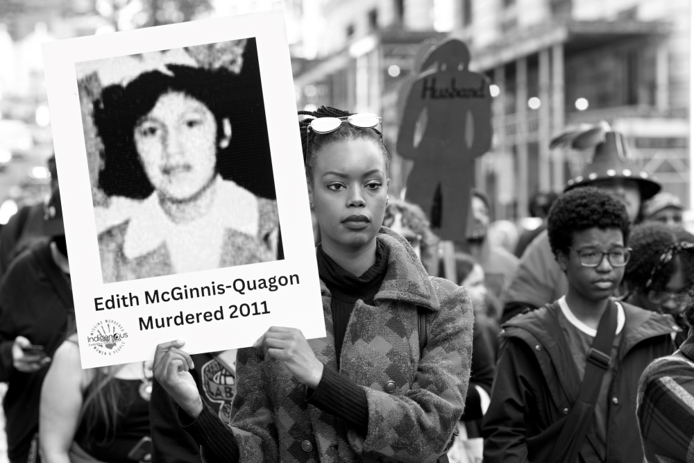 Young Black woman holds placard showing photograph of young girl with dark hair and wearing dress with Peter Pan collar.